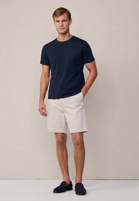Marineblå t-shirt med rund hals, beige shorts og mørkeblå loafers. Modellen står foran en neutral baggrund og viser en afslappet sommeroutfit.