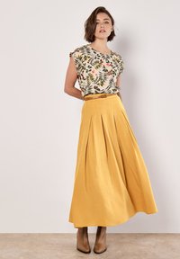 Top corto de flores con patrones verdes y naranjas, combinado con una falda larga amarilla con pliegues. Botas marrones de tacón acentúan el conjunto.