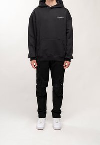 Felpa nera con cappuccio e tasca frontale, decorata con una grafica testuale; abbinata a pantaloni cargo neri con più tasche e sneakers bianche.