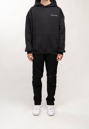 Schwarzer Hoodie mit einer Fronttasche und grafischem Text; kombiniert mit schwarzen Cargo-Hosen mit mehreren Taschen und weißen Sneakers.