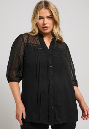 Blusa negra con mangas transparentes, lunares texturizados, pliegues debajo de la sisa, ribete de encaje en el escote y cierre de botones.