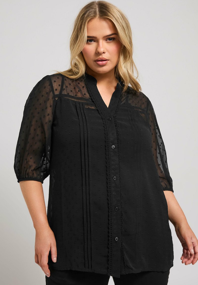 Svart blus med genomskinliga ärmar, texturerade prickar, veck under oket, spetsdetalj längs halsringningen och knäppning.