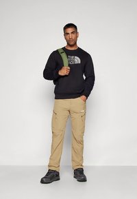 Fekete pulóver logóval, bézs cargo nadrág, szürke túracipők és zöld vállpánt. A modell semleges háttér előtt áll.