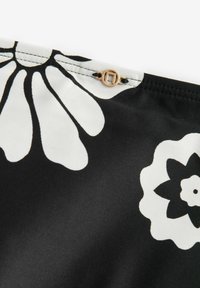 Tissu noir avec de grands motifs floraux blancs, comprenant un petit accent métallique doré et des détails de couture visibles le long du bord.