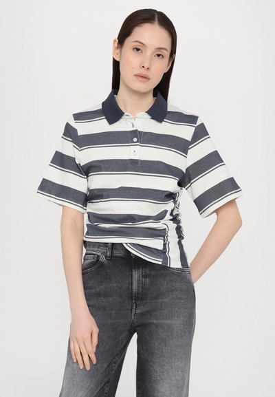 Polo skjorte med marineblå og hvite horisontale striper, korte ermer og marineblå krage. Brukt med mørkegrå jeans, ensfarget bakgrunn.