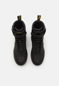 Botas de cuero negro hasta el tobillo con punteras redondeadas, cordones reforzados, una correa y una etiqueta de marca amarilla en la lengüeta. Acabado suave, diseño robusto.