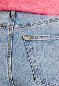 Helle blaue Denim-Jeans mit orangefarbenen Nähten, ausgestattet mit einer Gesäßtasche und einer weichen pinken Stricktextur oben.