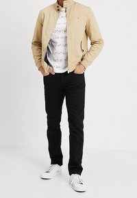 Vaaleanbeige vetoketjullinen takki, jossa on pystykaulus, yhdistettynä valkoiseen kuviolliseen paitaan, mustiin housuihin ja valkoisiin lenkkareihin. Yksinkertainen muotoilu, rento tyyli.