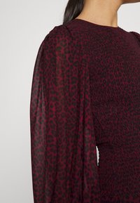 Vestito bordeaux con maniche a sbuffo trasparenti caratterizzate da una stampa leopardata nera. Il corpo del vestito ha dettagli arricciati, creando un effetto testurizzato.