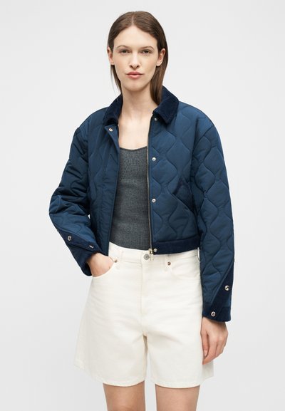 Jeune femme portant une veste matelassée bleu marine sur un haut gris sans manches et un short blanc taille haute, debout avec une main dans la poche.