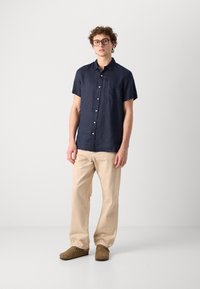 Korte mouwen marineblauw linnen shirt met een borstzak, gecombineerd met beige losse broek en zachte bruine instappers.