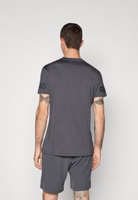 Mann mit Schmetterlingstattoos auf Armen und Beinen, der ein dunkelgraues T-Shirt und Shorts trägt und vor einem schlichten hellen Hintergrund steht.