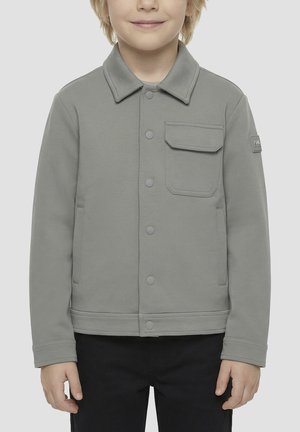 Enfant portant une veste grise boutonnée avec une poche à rabat sur le devant et deux poches latérales, associée à un pantalon noir.