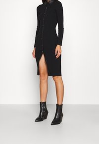 Svart långärmad klänning med knapplist fram och krage, som har en sidöppning och texturerad tyg. Matchad med svarta ankelskor.
