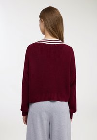 Donna con lunghi capelli castani indossa un maglione lavorato a maglia di colore marrone scuro con colletto a righe bianche e pantaloni grigio chiaro, con la schiena rivolta verso la fotocamera.