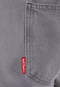 Gray denim fabric features a side pocket; a small red tag with "DADA" is visible, indicating the brand.