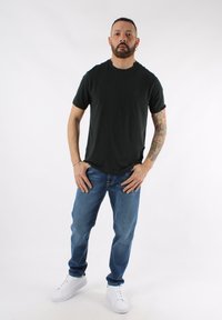 Camiseta negra de manga corta, vaqueros azules y zapatillas blancas. La camiseta tiene un escote redondo. Los vaqueros presentan un corte clásico y un lavado claro.