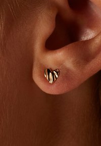 Boucle d'oreille en or en forme de cœur avec une surface texturée et ondulée, accrochée à un lobe, mettant en avant sa finition brillante et son design compact.