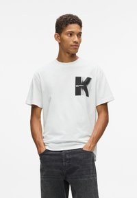 Jauns vīrietis, tērpies baltā T-kreklā ar melnu stilizētu "K" grafiku un melnās džinsa biksēs, stāv ar rokām kabatās, raugoties uz sāniem.