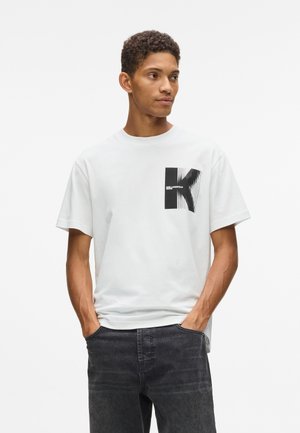 Mladý muž v bielom tričku s čiernym stilizovaným grafickým symbolom "K" a čiernych džínsach, stojaci so založenými rukami v kapsách a pozerajúci sa do strany.