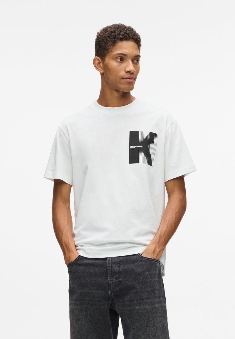 Jauns vīrietis, tērpies baltā T-kreklā ar melnu stilizētu "K" grafiku un melnās džinsa biksēs, stāv ar rokām kabatās, raugoties uz sāniem.