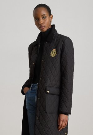 Donna che indossa un lungo cappotto trapuntato nero con emblema dorato, dolcevita nero e jeans blu, in piedi davanti a uno sfondo grigio uniforme.