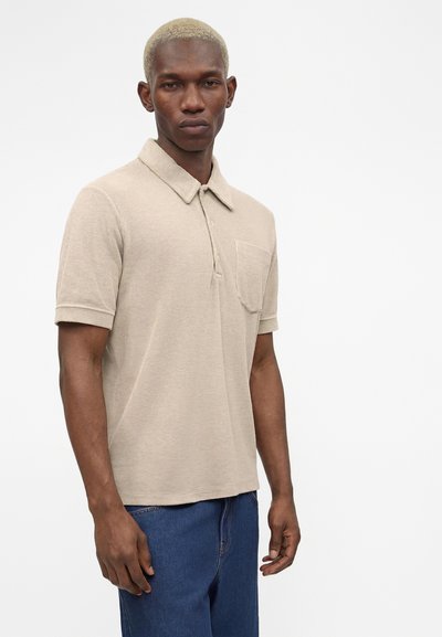 Άνδρας με κοντά ξανθά μαλλιά που φοράει μπεζ κοντομάνικο πόλο και μπλε τζιν μπροστά σε απλό λευκό φόντο.