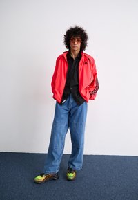 Red athletic jacket with black stripes, black shirt, loose denim pants, and multicolored sneakers with a textured sole.