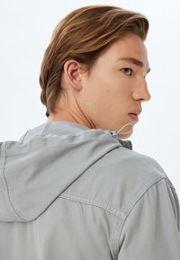Chaqueta con capucha de color gris claro y textura suave, con cierre de cremallera y cordón ajustable. El diseño incluye yugo trasero y detalles de costura.