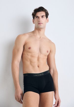 Oberkörperfreier Mann mit durchtrainiertem Körper, der schwarze Lacoste-Boxershorts trägt, vor einem schlichten weißen Hintergrund steht und leicht nach oben blickt.
