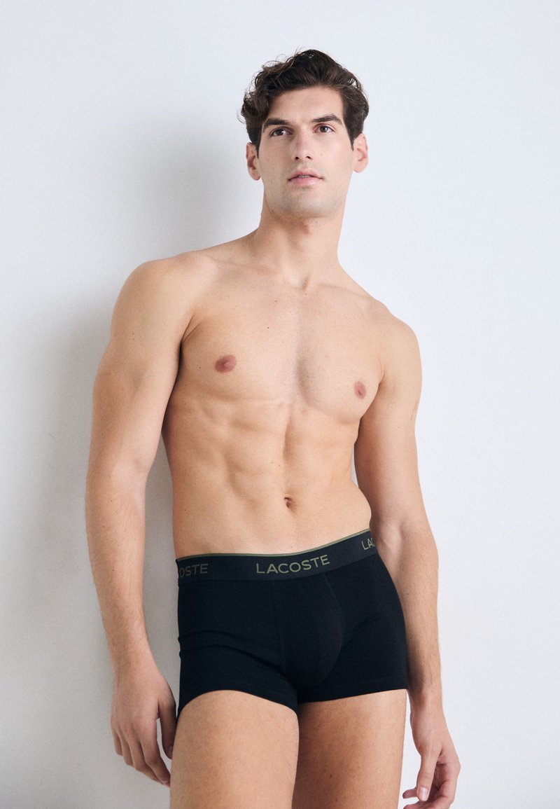 Homem sem camisa com um físico tonificado, vestindo boxers pretos da Lacoste, de pé contra um fundo branco liso, olhando ligeiramente para cima.