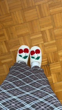 Pantofole bianche pelose con ricami di ciliegie rosse e foglie verdi; abbinate a una gonna a fantasia quadrata; sfondo di pavimento in legno.