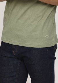 Homme portant un t-shirt vert olive avec un petit détail en losange blanc et un jean en denim bleu foncé, les bras croisés au-dessus de la taille.