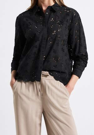 Kobieta nosząca czarną kwiecistą koszulę z dziurkami i guzikami oraz beżowe spodnie ze sznurkiem, ręce w kieszeniach, na jednolitym jasnym tle.