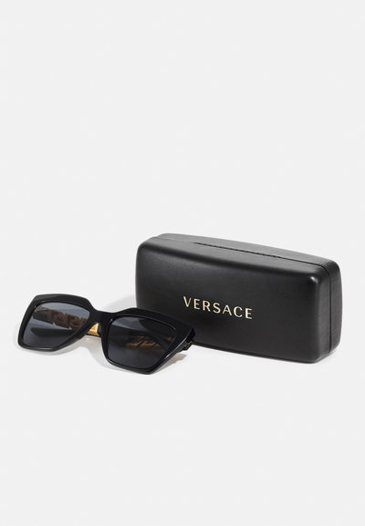 Óculos de sol quadrados pretos com armação grossa mate e lentes tonais, acompanhados de uma caixa preta da Versace com logótipo dourado.
