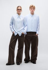 Ανοιχτό μπλε πουκάμισα με μεγάλους μυτερούς γιακάδες, σε συνδυασμό με φαρδιά σκούρα καφέ παντελόνια. Τα πουκάμισα έχουν εμφανείς λεπτομέρειες κουμπιών και υφή υφάσματος.