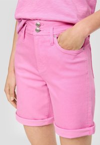 Primer plano de una persona que lleva puestos pantalones cortos de dobladillo enrollado de color rosa brillante y una camisa rosa a juego, con una mano en el bolsillo delantero.