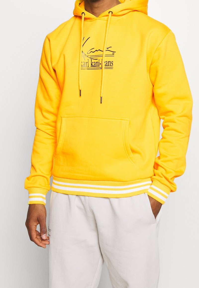 Sudadera amarilla hecha de tejido suave, con un bolsillo tipo canguro, puños y dobladillo de rayas blancas acanaladas, y un logo impreso en el frente.
