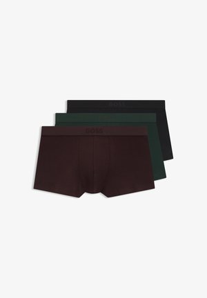 Homme en forme à la peau foncée portant un boxer marron foncé uni, debout devant un fond clair uni.