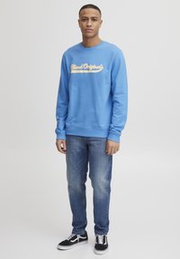 Sudadera azul clara con el logo "Blend Originals" en amarillo, combinada con jeans azules y zapatillas en blanco y negro. Textura suave, corte casual.