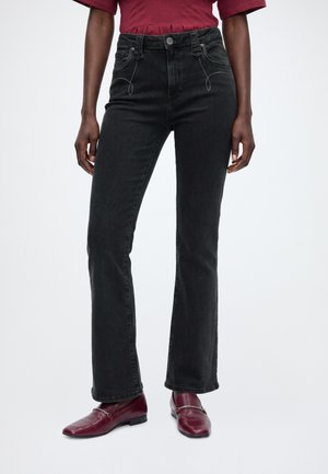 Person iført højtaljede sorte jeans, bourgogne læder loafers med kædedetalje og en bourgogne kortærmet top, stående på hvid baggrund.