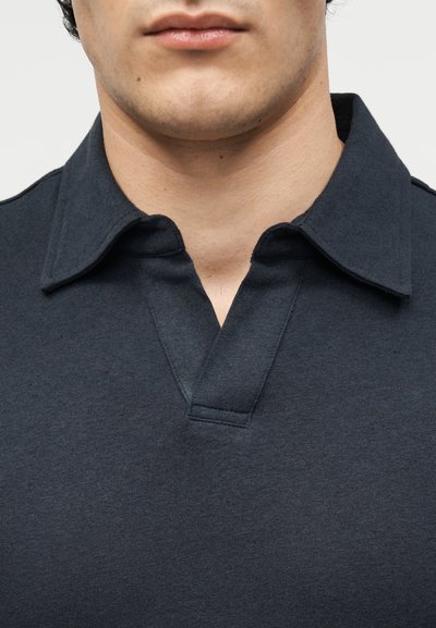 Κοντινό ενός άνδρα που φοράει σκουρόγκρινο πόλο με ανοιχτό γιακά και χωρίς ορατά κουμπιά.