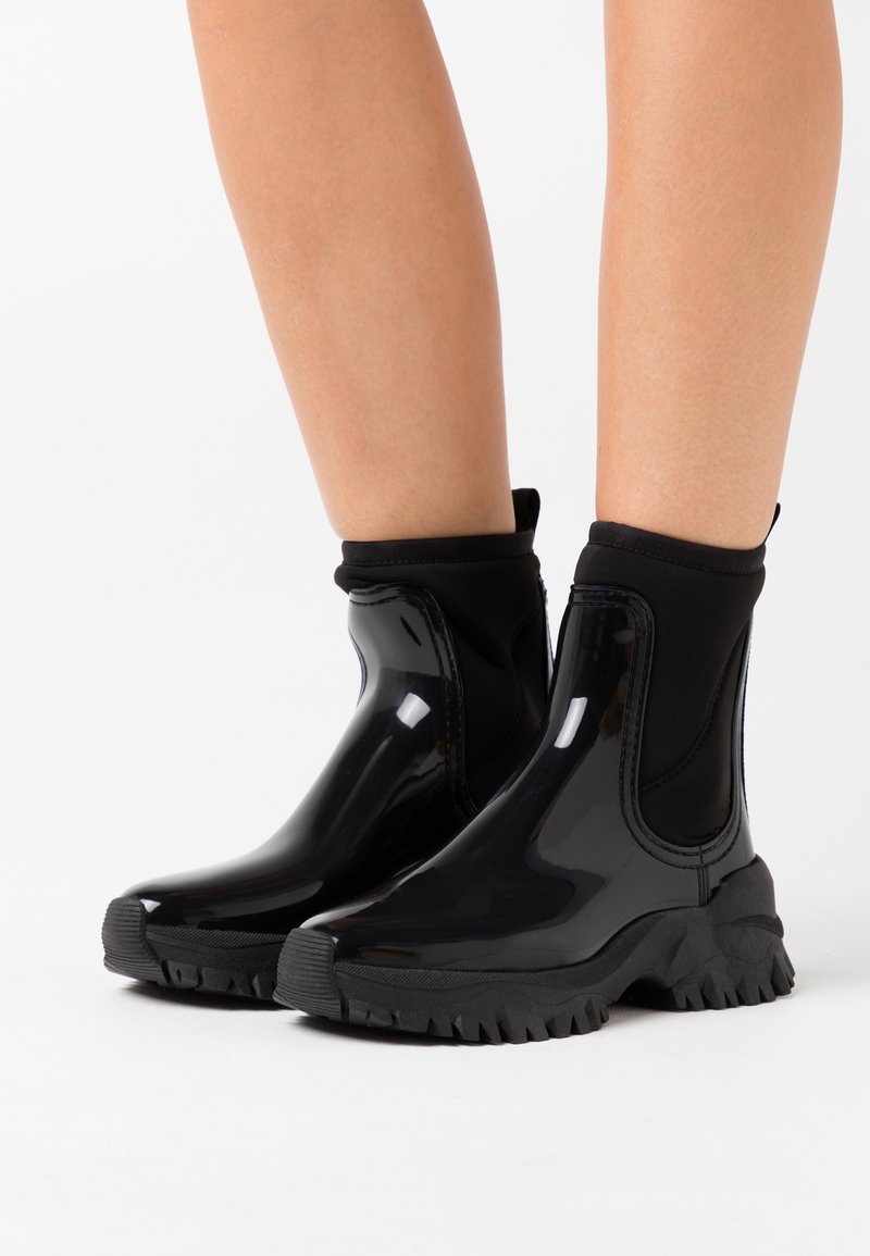 Bottes noires à cheville avec une finition brillante, tige élastique semblable à une chaussette, et semelle en caoutchouc épaisse avec un motif de bande de roulement texturé.