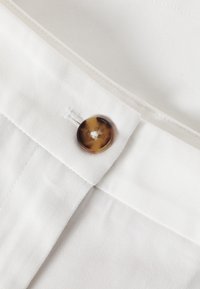 Tessuto bianco con una superficie liscia e un bottone marrone marmorizzato. Sono visibili cuciture da vicino vicino al bottone e al bordo del tessuto.