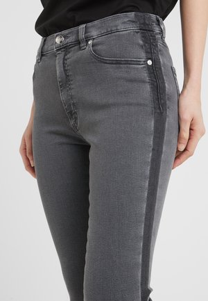 Jeans de denim gris con un diseño ajustado, que presentan una franja lateral vertical más oscura, bolsillos frontales y acentos en metal plateado. Textura suave.