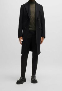 Osoba ubrana w czarny, podwójnie zapinany długi płaszcz, oliwkową golf, czarne spodnie i czarne, masywne botki, stojąca na szarym tle.