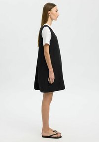 Zwarte mouwloze jurk van lichte stof, gedragen over een wit T-shirt met korte mouwen, gecombineerd met zwarte slippers. Eenvoudige A-lijn silhouet.