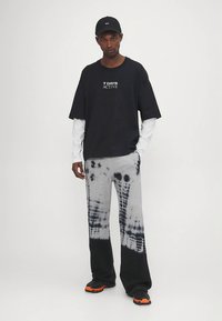 Juoda grafikos marškinėliai su baltais užrašais, pilkos tie-dye kelnės su juodais akcentais ir juodi sportbačiai su oranžiniais padais.