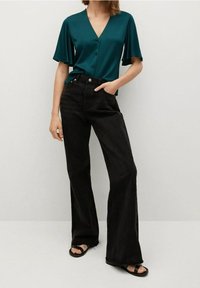 Blouse en satin turquoise avec manches volantes, col en V et boutons, associée à un jean noir évasé taille haute et des sandales plates noires.