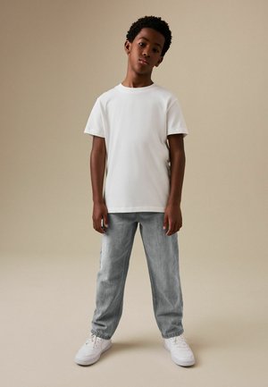 Junge steht mit neutralem Gesichtsausdruck, trägt ein weißes T-Shirt, hellgraue Jeans und weiße Turnschuhe vor einem schlichten beigen Hintergrund.